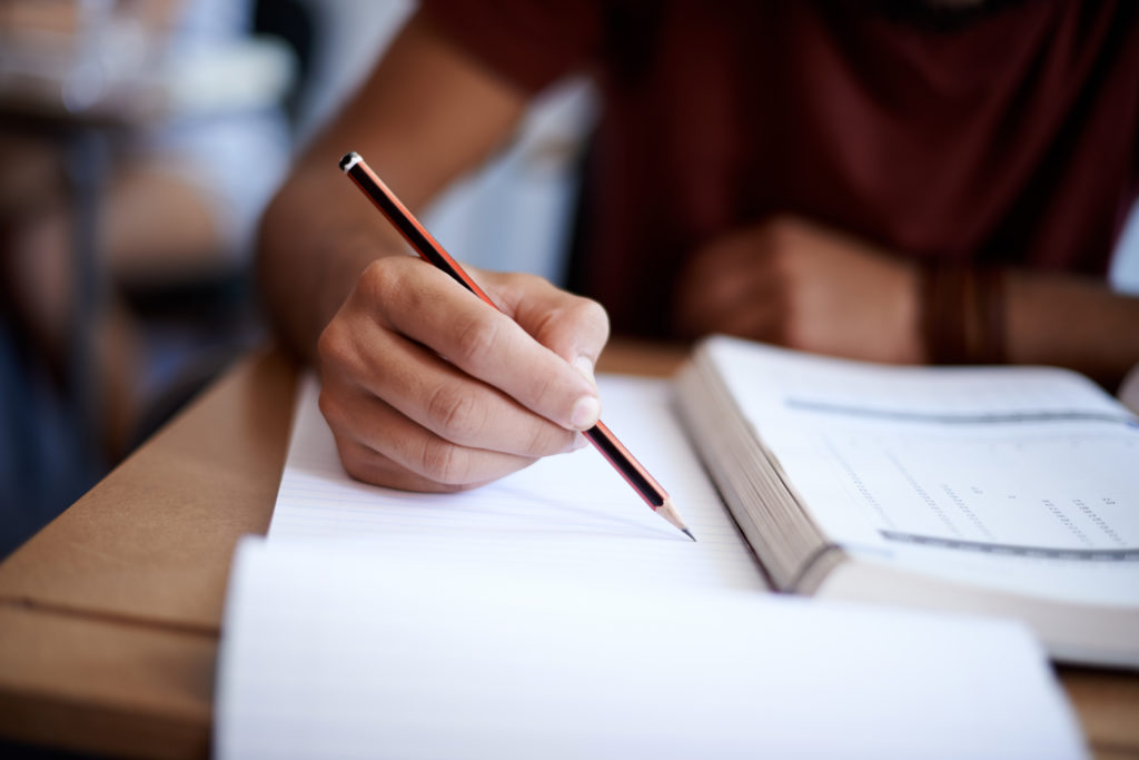 A student holds a pencil, with open textbook and note paper.