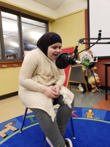 A student sits on a stool and speaks into a microphone to record a podcast.