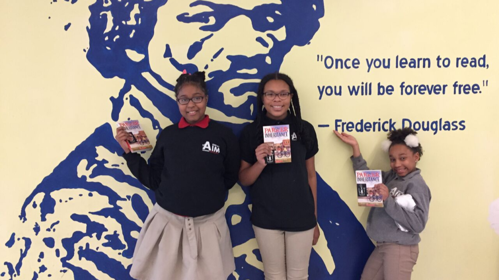 Three students stand in front of a wall painted with Frederick Douglass' image and a quote. Each student holds a book.