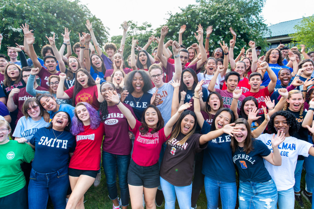 Dozens of students in college shirts cheer together.
