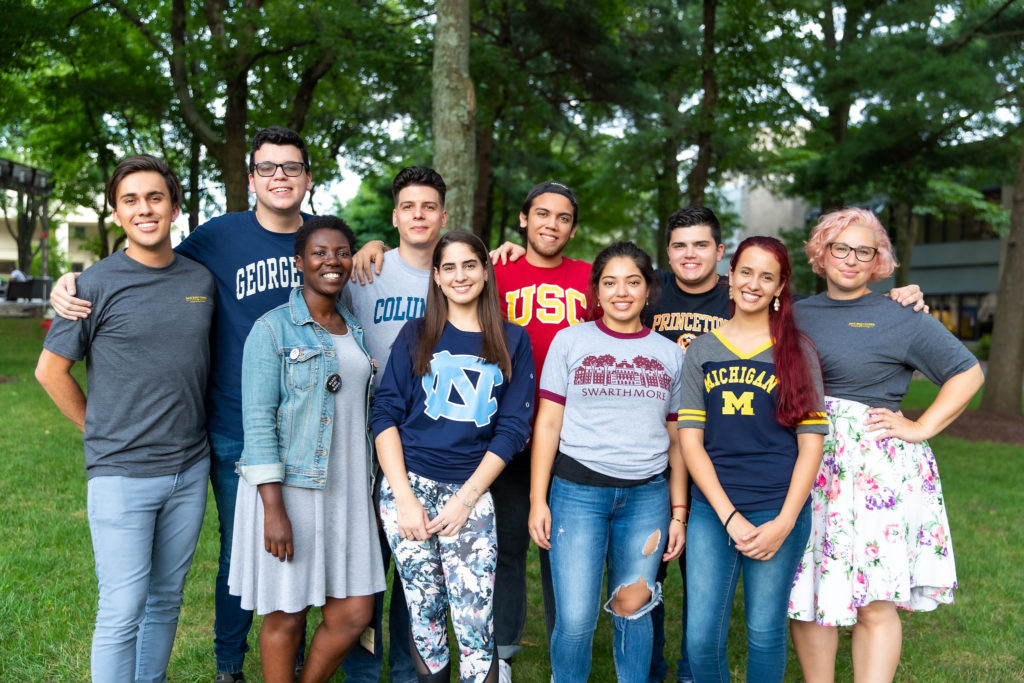 10 students stand together outside, each wearing a t-shirt from a different college or university.