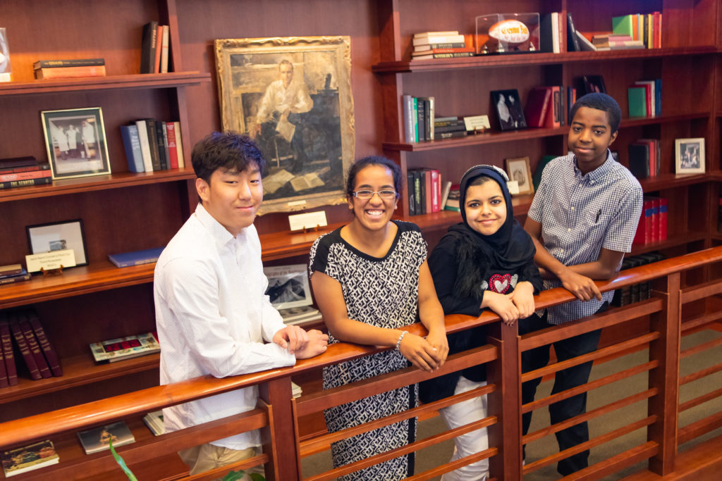 Four students stand in front of a large bookcase.