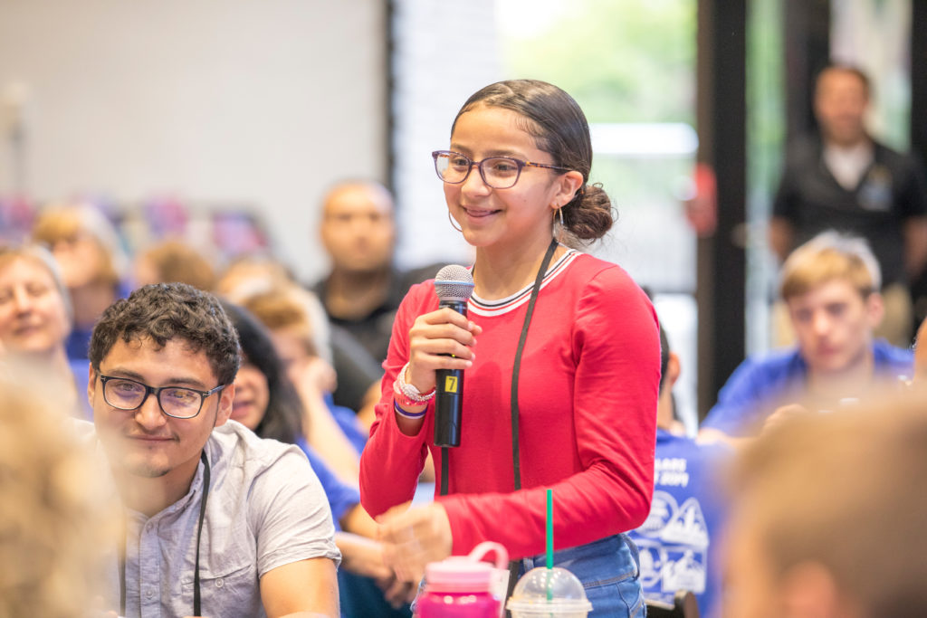 A girl holding a microphone is standing up amongst an audience of seated students.