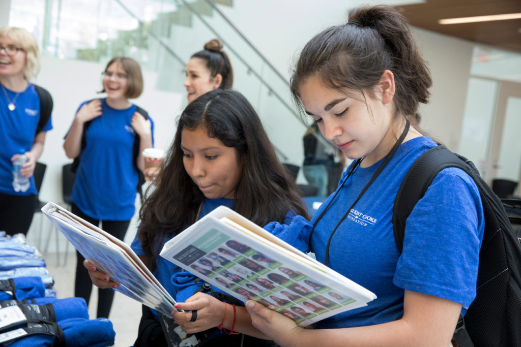 Two young female students look down at binders. They are inside a building on college campus with other students.