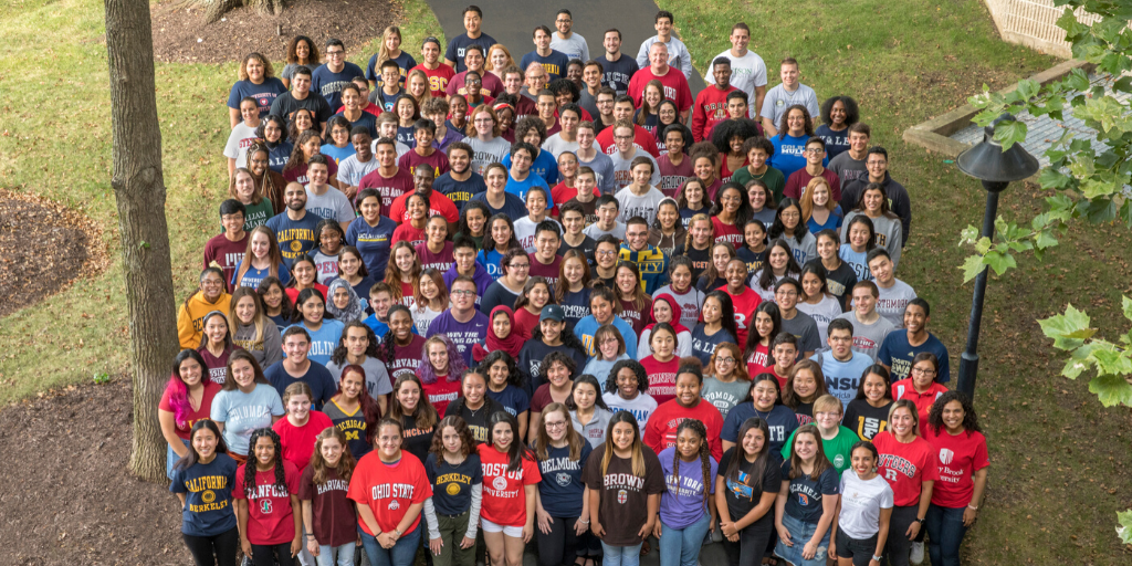 A large group of students stands outside wearing college t-shirts.