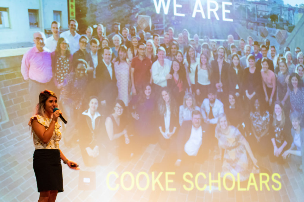Woman holding a microphone in front of a photo backdrop of a group of students